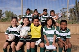 A group of mainly Indigenous children in sports clothes sit smiling at the camera with red dirt behind them.