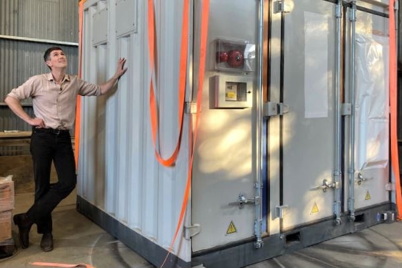 Indigo Power director Ben McGowan stands next to a large square, a community battery.