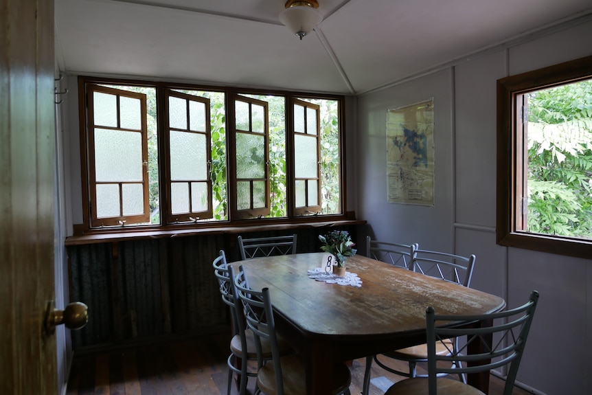 Una mesa y sillas de madera dentro de una pequeña habitación con ventanas abiertas. 