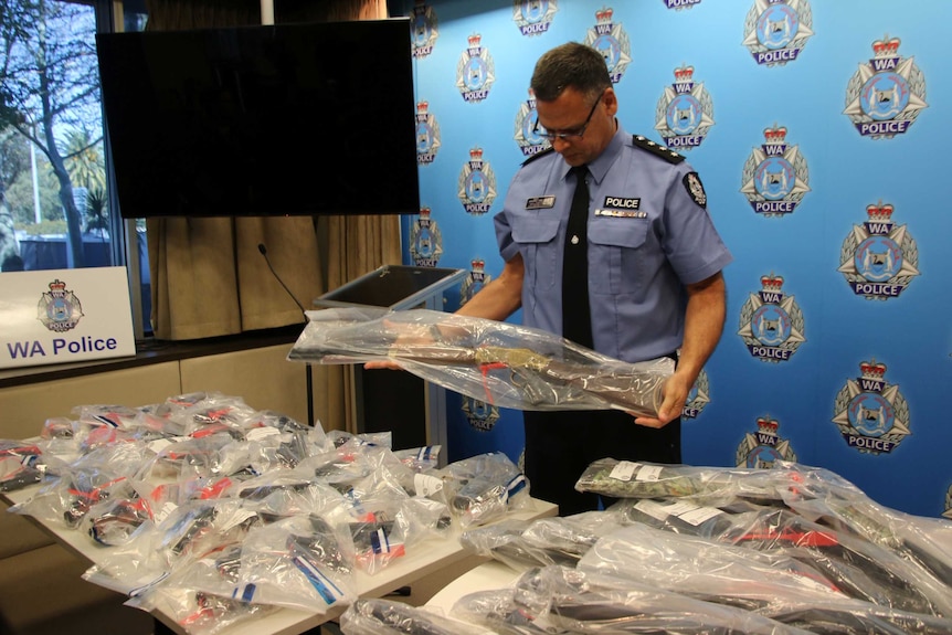 A police officer stands at a table covered with guns in plastic bags holding a rifle in a plastic bag.