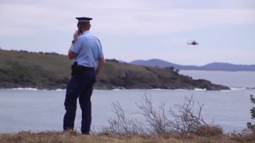 A policeman in short-sleeves stands on a hill overlooking the sea as a helicopter flies low over rocky coast in the distance