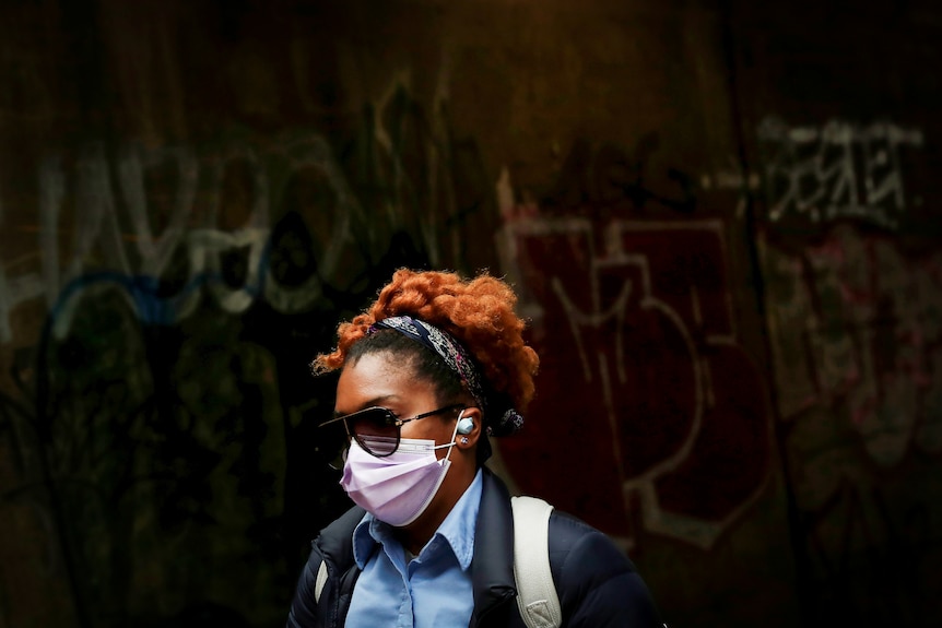 A woman in a purple surgical face mask walks past a graffiti wall 