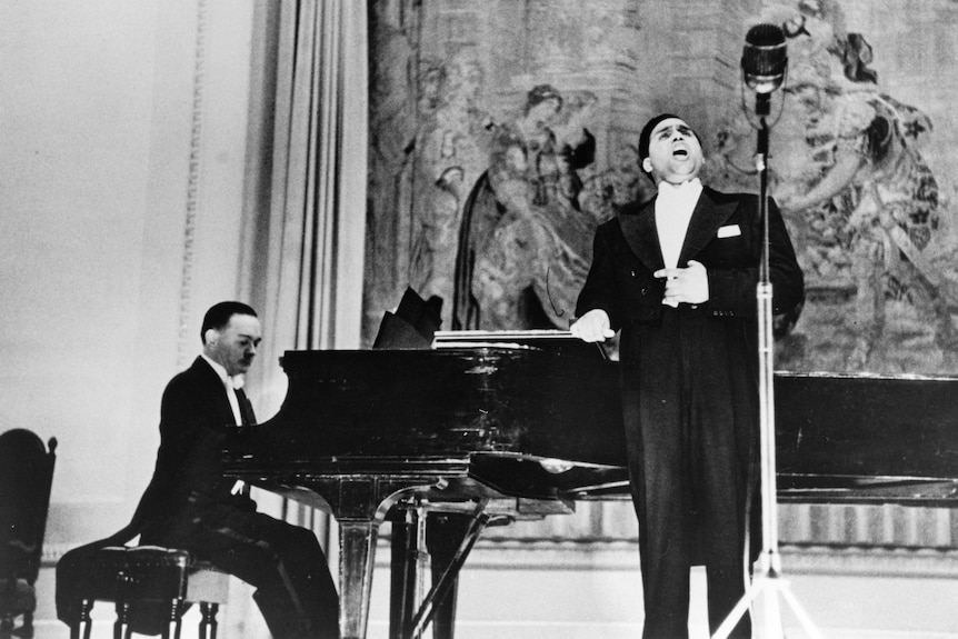 A man wearing a suit sings in front of a large microphone while a man sits playing a grand piano.