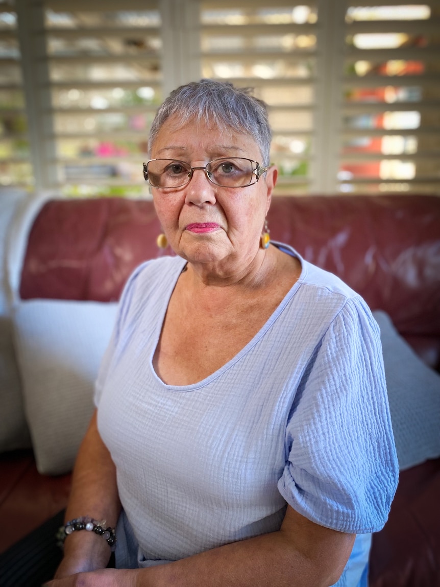 Doris looks unhappy, sitting on a couch in her home. She has short, grey hair, glasses and is wearing a pale blue top.