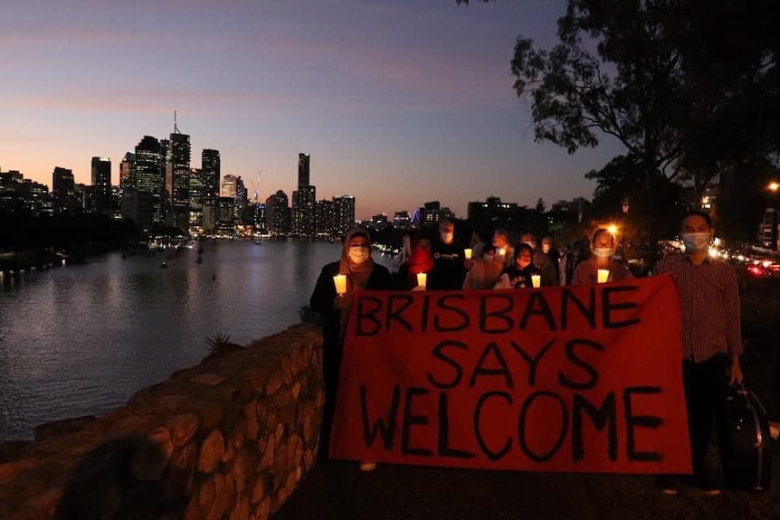 People stand holding candles and a sign that says "Brisbane says welcome" with the Brisbane skyline behind them.