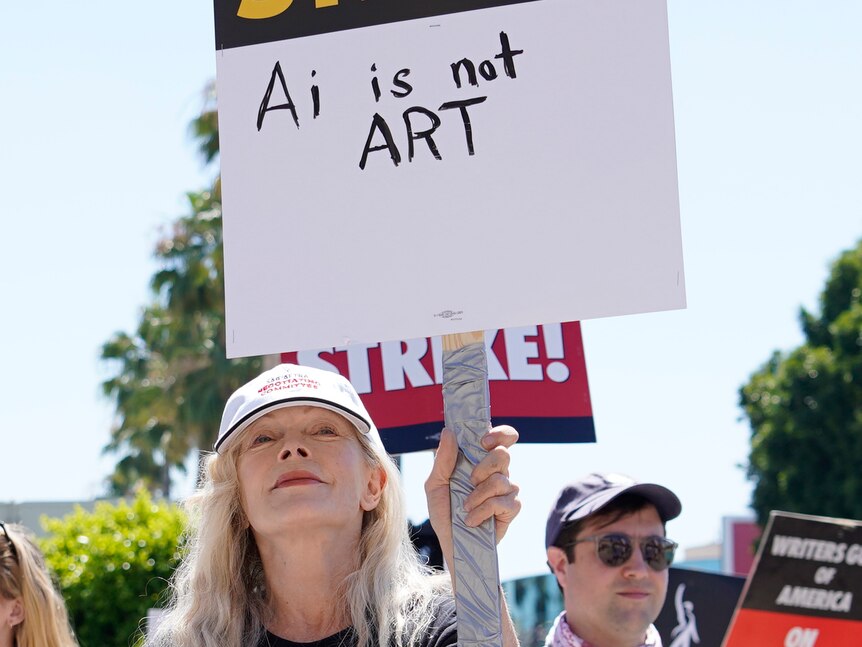A woman with blonde hair holds a sign saying 'AI is not art' 