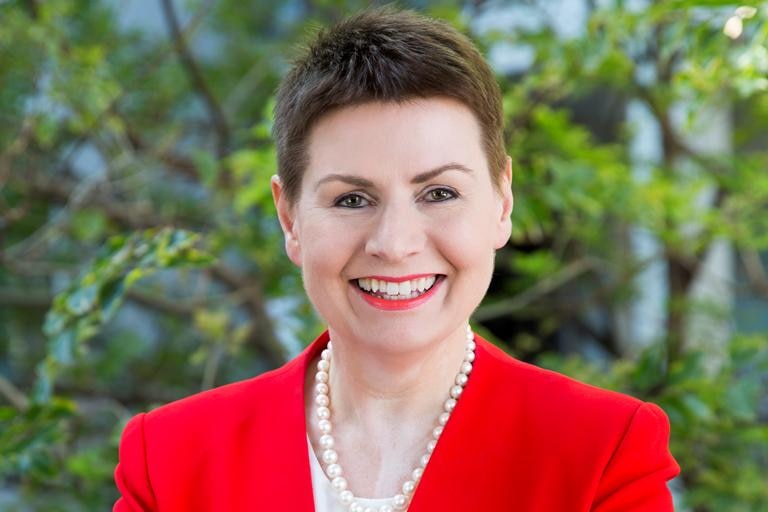 A woman with short brown hair, in a bright red formal jacket, with arms crossed, smiling.