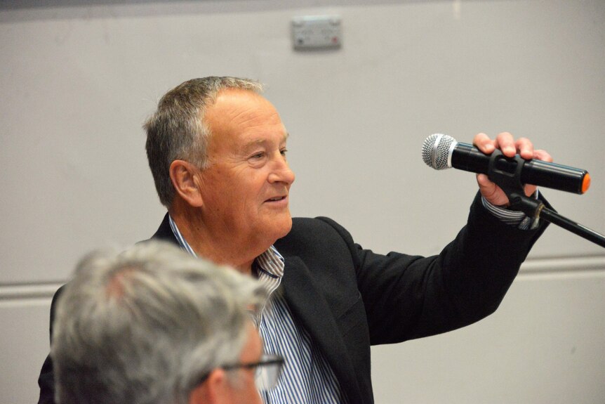 An older man in a suit reaches out to a microphone while looking off camera
