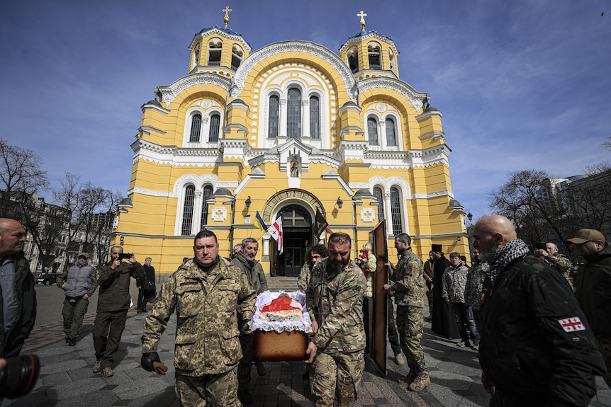 Procesión sombría de hombres que llevan un ataúd blanco y rojo cubierto de banderas con una iglesia amarilla y blanca detrás en un día soleado