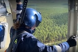 A person leans out the side of a helicopter looking over vast green forest