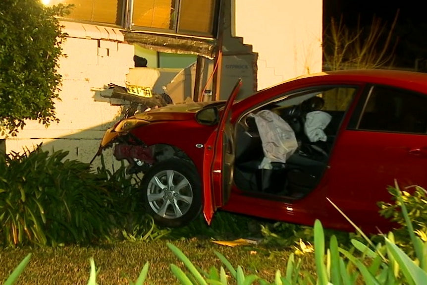 A damaged car sits against a house wall with a large hole in it.