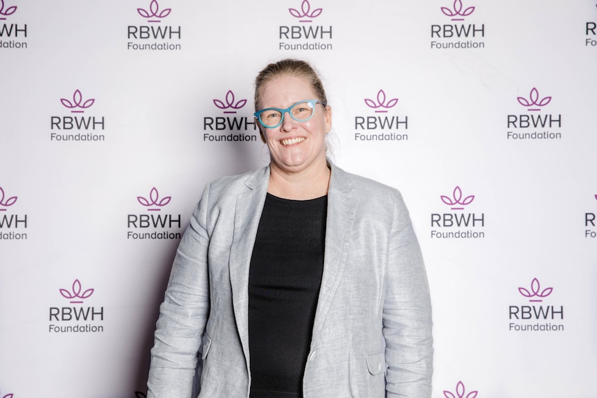 A woman in blue glasses smiles in front of a RBWH wall.
