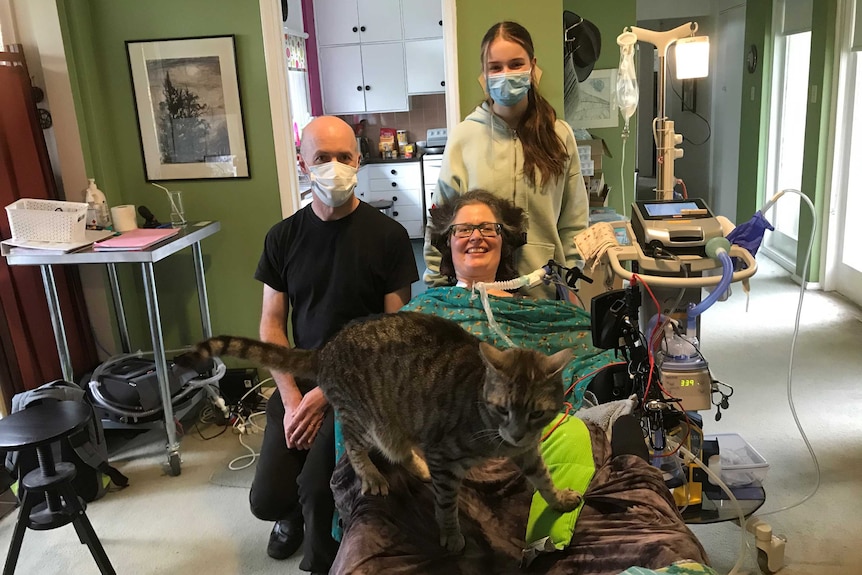 A woman is on a medical bed attached to equipment and a man and a girl stand beside her in medical masks.