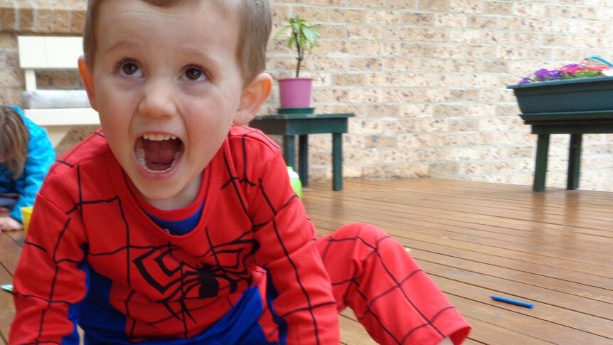A young boy in a spiderman suit opens his mouth excitedly, facing just off camera.