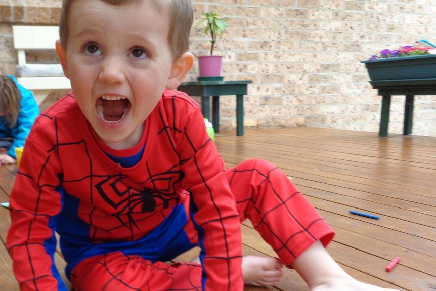 A young boy in a spiderman suit opens his mouth excitedly, facing just off camera.