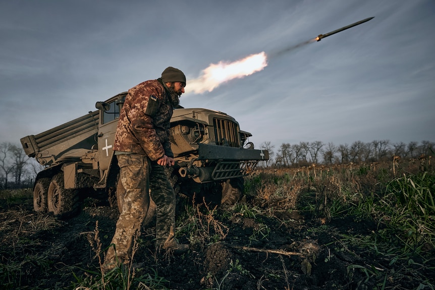 A truck with a rocket laucher system mounted to the back fires a rocket as a soldier stands nearby.