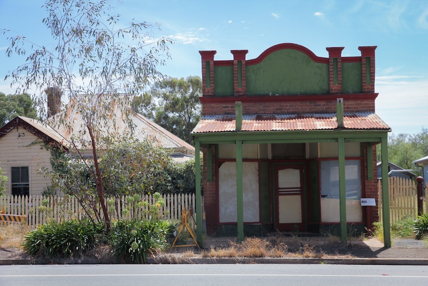 A green building, which appears to be in quite poor condition, on the side of the road in a country town.