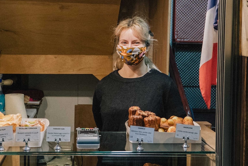 A smiling woman wearing a patterned mask stands behind a display case full of pastries.