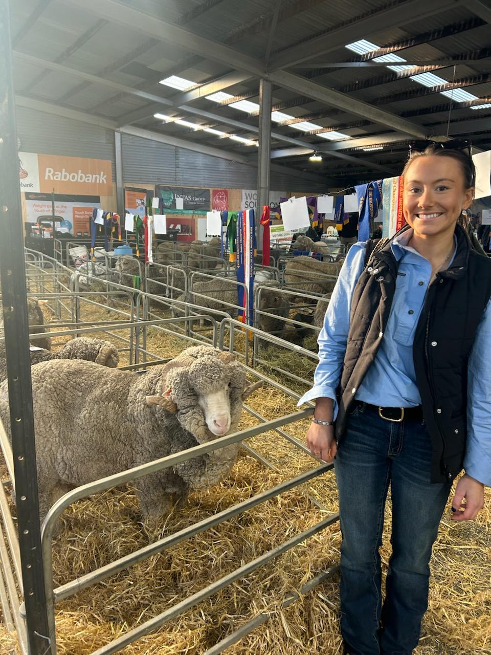 A woman stands smiling next to a sheep in a pen inside.