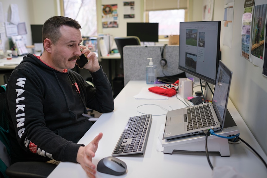 Mark sits at his desk with a screen and laptop open in front on him talking to someone on his mobile phone.