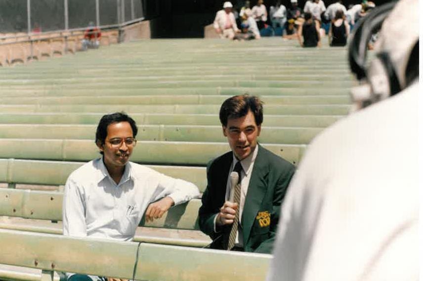 Gee holding microphone sitting next to Bhogle while cameraman filming.