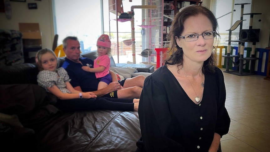 Woman with glasses looks ahead, while man and two young children sit on couch in the background.
