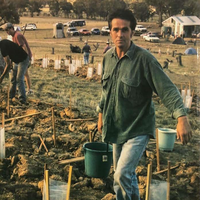 a man holds a bucket of water, behind him are tree guards and people working in a paddock