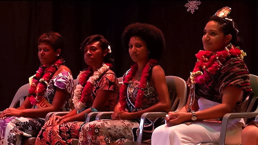 Four women in traditional clothing sit on chairs on a stage.