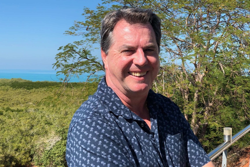 Image of a man standing at a fence in Broome, with a bay and trees in the background.