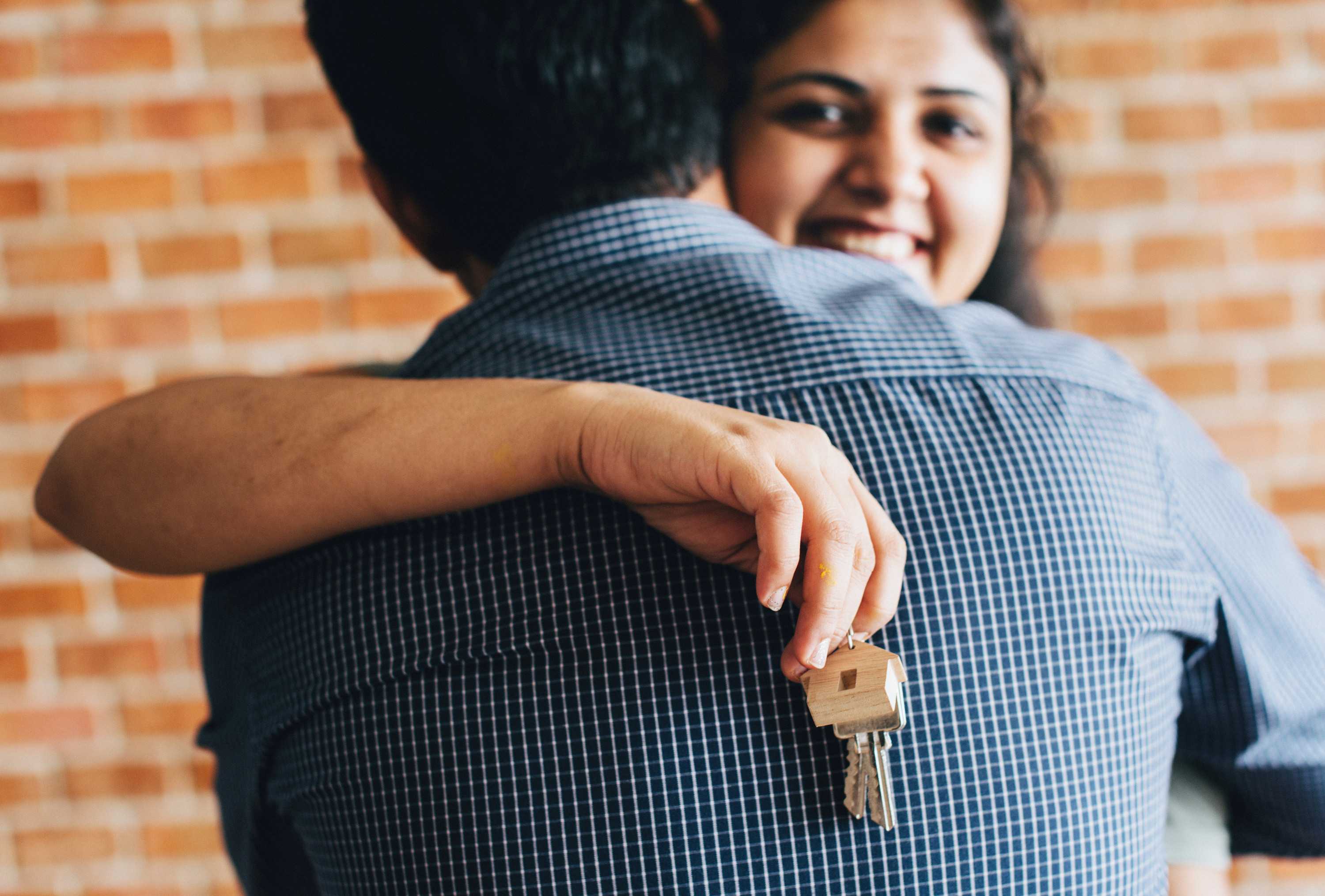 A smiling woman hugs a man while holding a keys on a keyring with a wooden house.