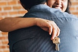A smiling woman hugs a man while holding a keys on a keyring with a wooden house.