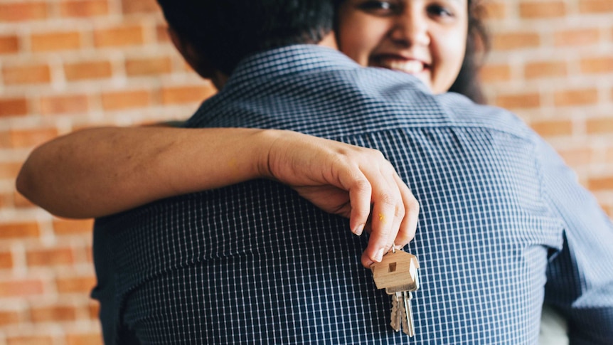 A smiling woman hugs a man while holding a keys on a keyring with a wooden house.