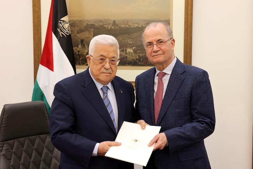 Two mean wearing suits standing next to each other holding a piece of paper with a palestinian flag behind them