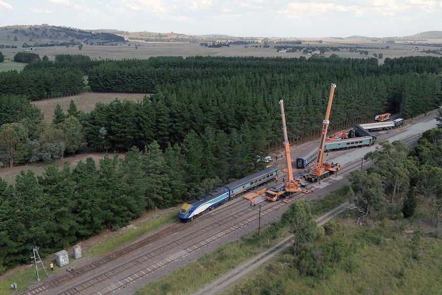 A tall crane stands alongside a derailed train with a number of workers in reflective vests.