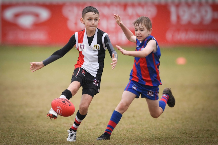 A junior Aussie rules player gets a kick off as another player gives chase in the rain.