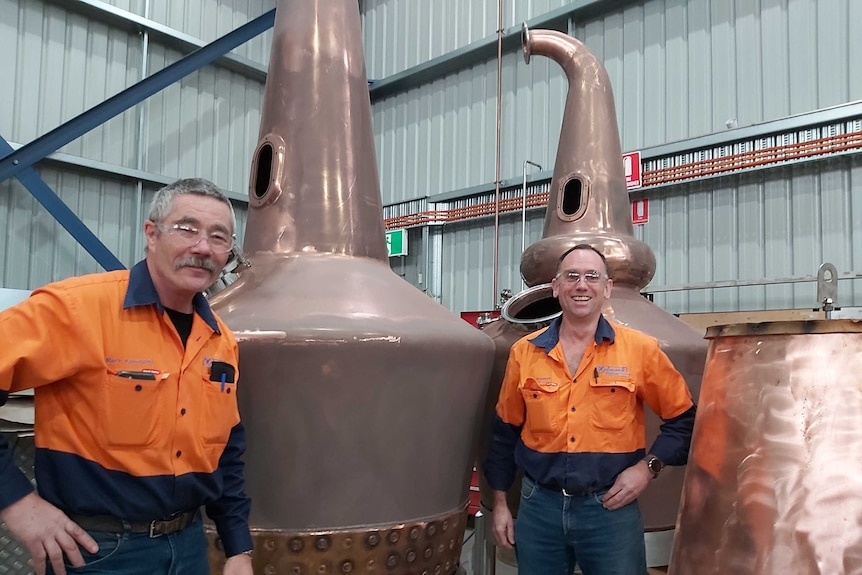 two men stand in front of two large copper pot whisky stills inside a steel fabrication shed