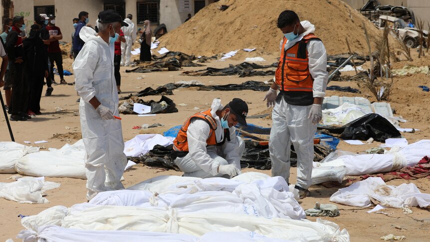 Workers in forensic overalls examine a row of shrouded corpses.