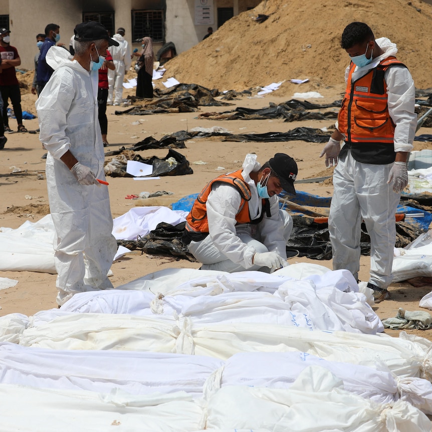 Workers in forensic overalls examine a row of shrouded corpses.