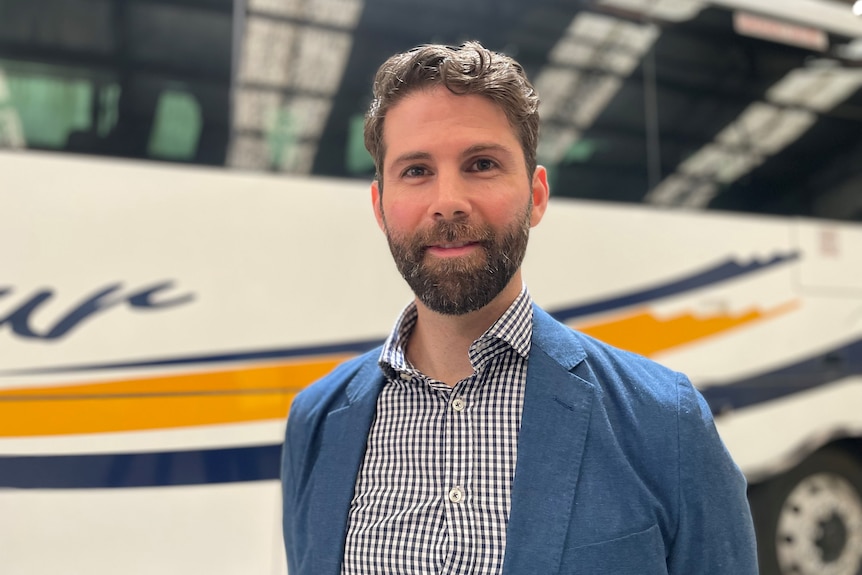 A man with a short beard, wearing a blue blazer, stands in front of a bus