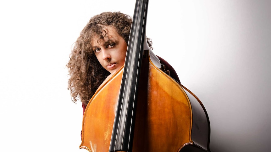 A photo of Helen hiding behind her upright double bass. Just her head is poking out. She has curly brown hair.