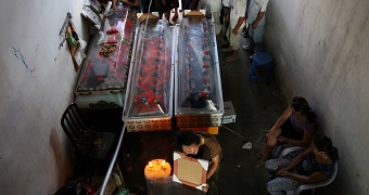A group of people standing behind three coffins. A woman sits in front of the coffins holds a photo, mourning.
