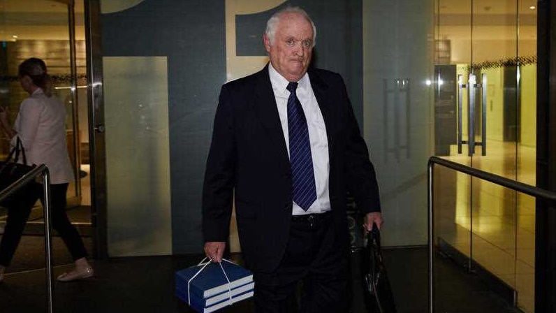 A man with white hair, wearing a suit, carried three hardcover books in one hand.