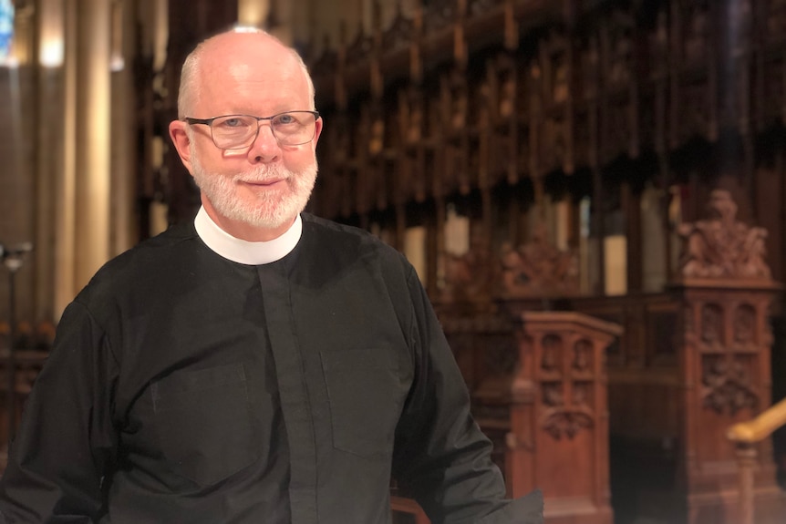 A reverend with white hair, a beard and glasses stands in a church