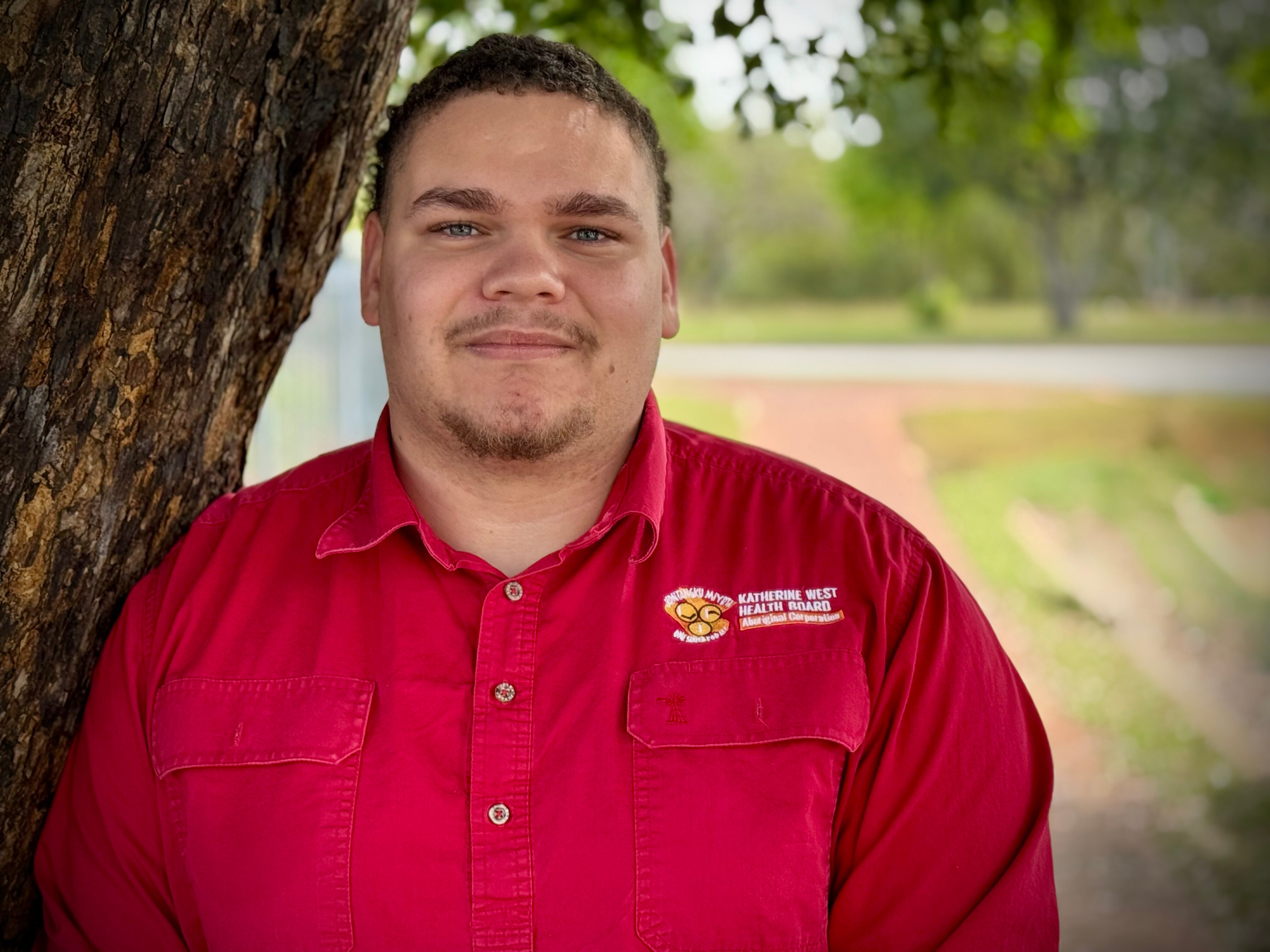 A First Nations young man in a red tshirt, short brown hair and goatie, smiling at camera. Standing next to a tree