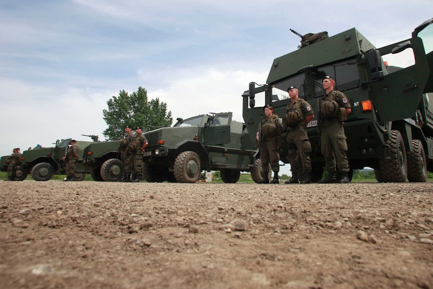 Soldiers stand in front of military vehicles.