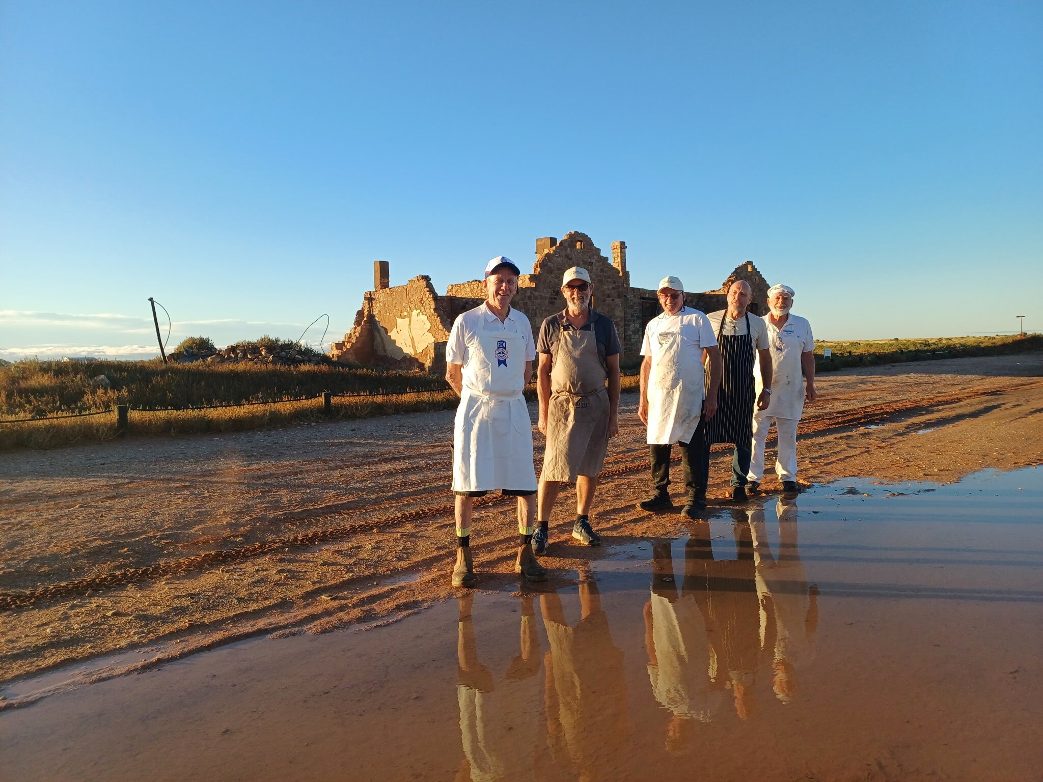 Five smiling men in aprons stand in front of the ruins of a stone building, on a clear outback day.