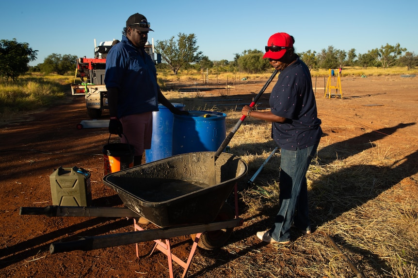 A photo showing two people mixing concrete in a wheel barrow with building materials in background