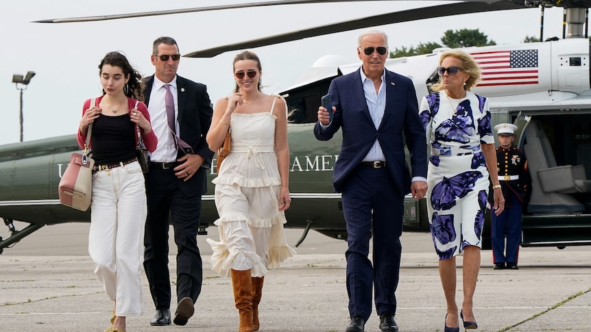 Two younger women, an older man and an older woman walking in front of a helicopter with a security guard in the background. 