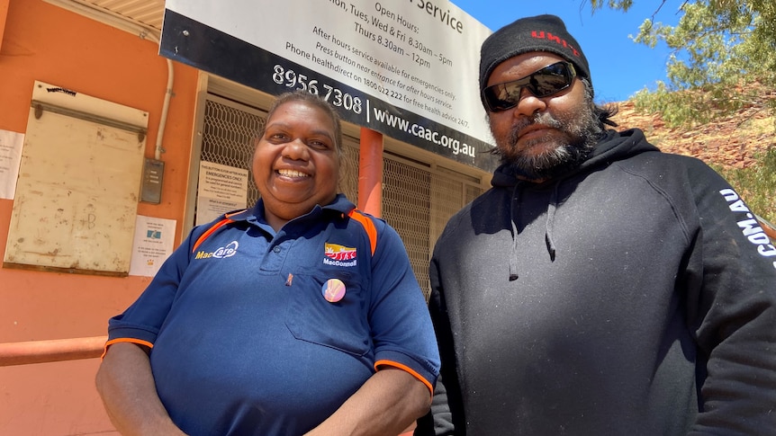 A woman wearing a blue shirt standing next to a man wearing a black hoodie and dark glasses out the front of an orange building.