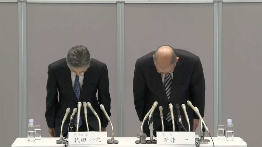 Two greying men in suits bow while stood behind a table during a press conference in Japan, with a grey wall behind them.